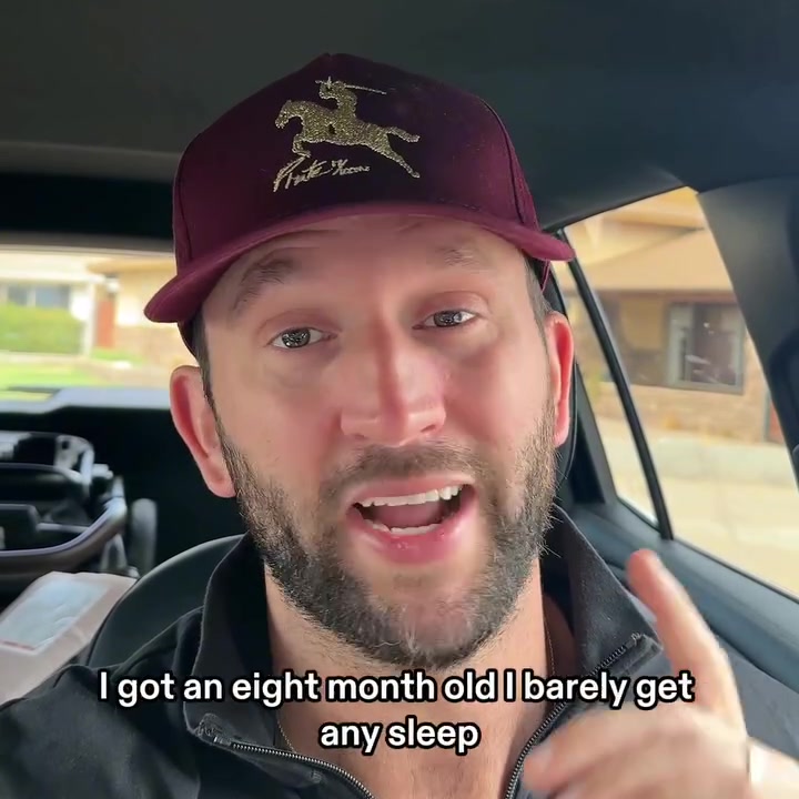 A bearded dad in a maroon cap and dark grey zip-up jacket filming himself in the driver's seat of a parked car, with the caption: I got an eight month old I barely get any sleep.